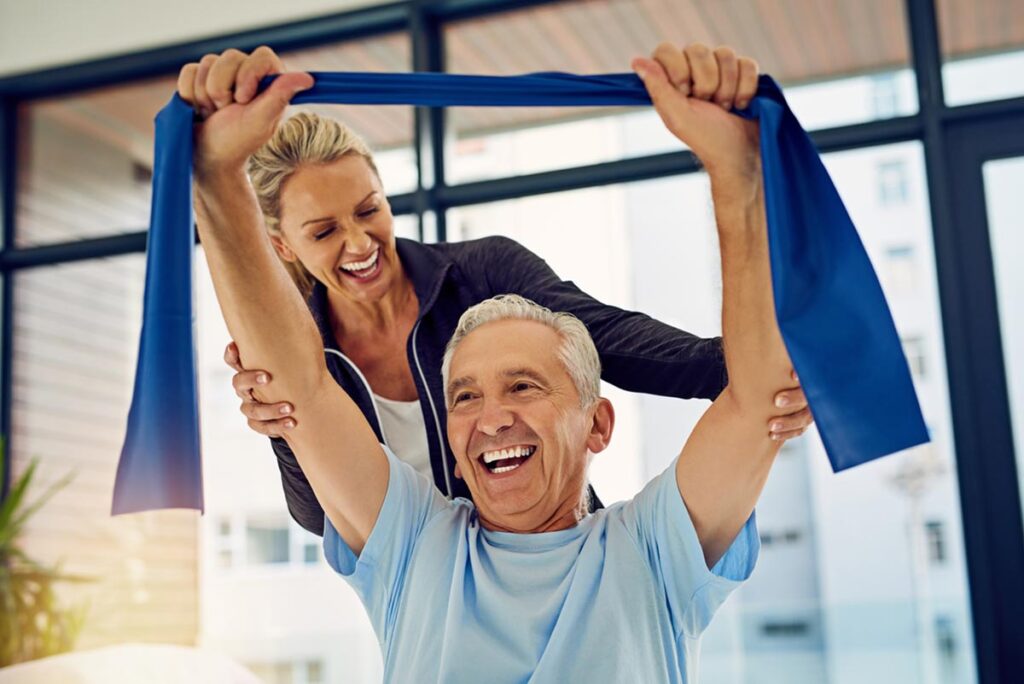 a senior in need of physical therapy holds a resistance band while doing exercises with his physical therapist