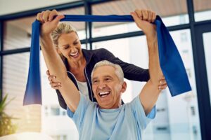 a senior in need of physical therapy holds a resistance band while doing exercises with his physical therapist